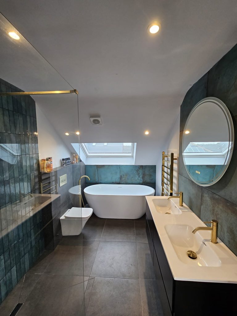 Freestanding bath beneath a rooflight, brass towel rail and double vanity with illuminated mirror in the loft en-suite